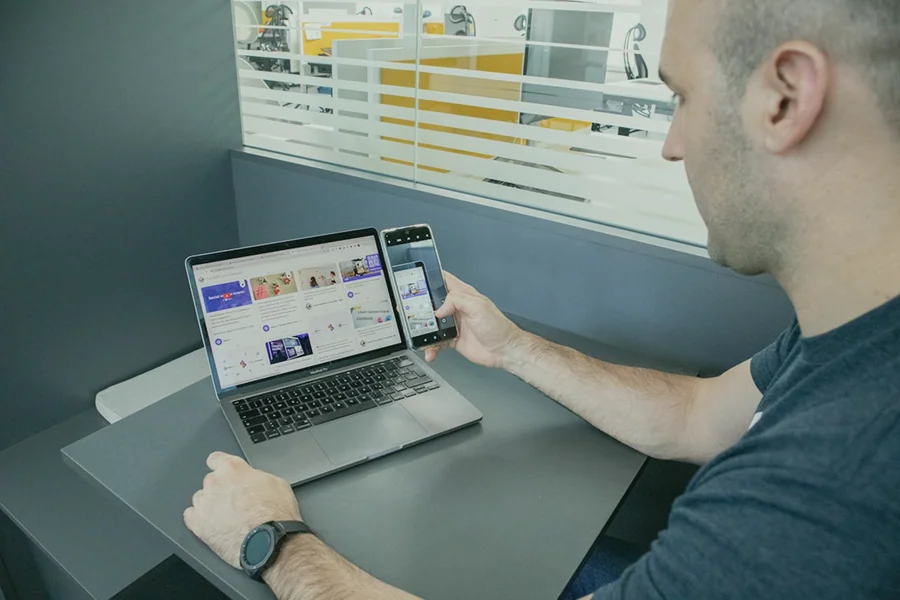 Un uomo è seduto alla scrivania con un computer portatile e uno smartphone nella mano destra. Entrambi i dispositivi visualizzano pagine web simili. Lo sfondo mostra pareti di vetro e attrezzature da ufficio.