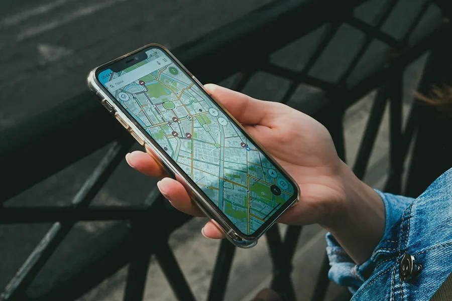 Una persona con in mano uno smartphone che visualizza una mappa digitale con diversi indicatori di posizione, in piedi accanto a una ringhiera di metallo nero all'aperto.