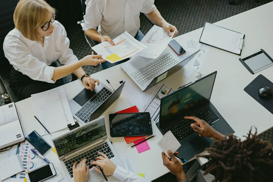 Quattro persone collaborano a un tavolo ingombro di computer portatili, fogli, grafici, note adesive e tablet, che sembrano discutere documenti e analizzare dati durante una riunione di lavoro.