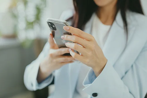 Una donna con un blazer azzurro tiene in mano e utilizza uno smartphone, con il volto parzialmente fuori dall'inquadratura e lo sfondo sfocato.