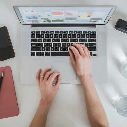 A person types on a laptop placed on a white desk with a smartphone, closed notebook, pen, water glass, and camera nearby. The laptop screen displays several image thumbnails.