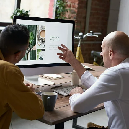 Two men sit at a wooden desk, looking at a computer screen displaying a design with text and images. One man points at the monitor, while the other listens. Coffee mugs, a notebook, and a keyboard are on the desk.