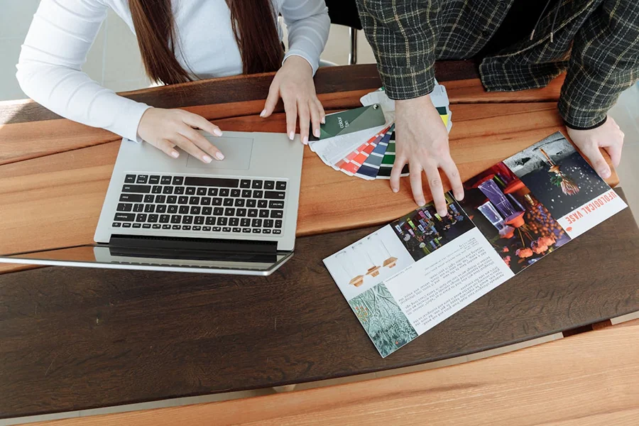 Due persone lavorano a un tavolo di legno; una utilizza un computer portatile mentre l'altra indica una brochure colorata e tiene in mano una serie di campioni di colore, suggerendo una discussione sul design o sui materiali di marketing.