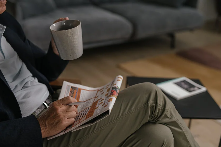 Una persona siede su un divano con in mano una grande tazza e legge un giornale. Su un tavolo vicino ci sono un libro chiuso e altri oggetti. L'ambiente sembra essere un accogliente salotto.